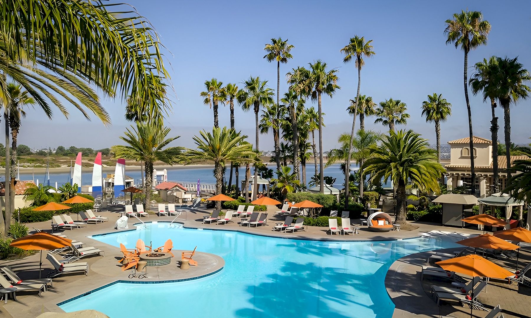 A sunny resort pool area with palm trees, lounge chairs, umbrellas, and people swimming and relaxing by the water, overlooking a calm view.