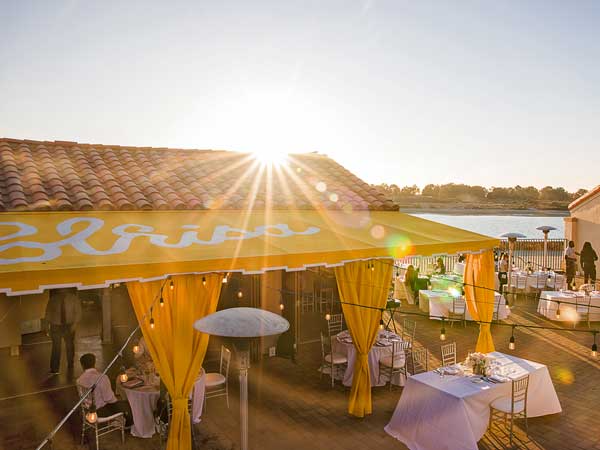 Sunlit outdoor beach party with yellow tents, string lights, cocktail tables, and a calm water backdrop at sunset, lively yet relaxed atmosphere.