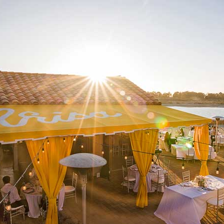A bright outdoor event at sunset with a yellow tent, string lights, and decorated tables by the water, guests mingling under canopies.