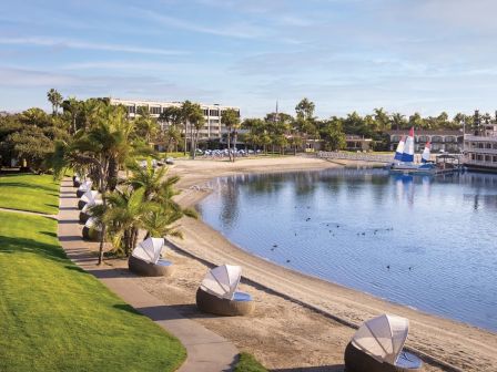 A calm coastal scene with a sandy beach, palm trees, and green lawns. Modern buildings line the shore, sailboats docked by a marina, and clear blue water reflecting a sunny sky.
