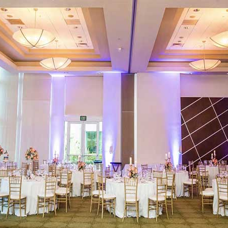 Elegant banquet hall set for a formal event with round tables, gold chairs, white linens, floral centerpieces, and warm purple lighting.