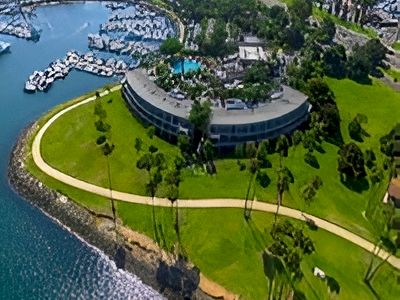 Aerial view of a curved modern building on a grassy cliff by the sea, with a winding path, palm trees, and a marina of boats nearby.
