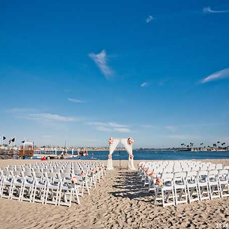 A sunny beach setup with rows of white chairs facing a floral arch over sandy shore, blue sky above and calm sea beyond.