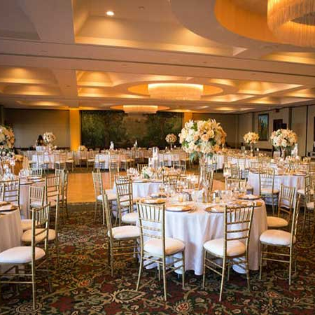A banquet hall set for a formal event with round tables, white linens, elegant floral centerpieces, golden chairs, and soft lighting on a patterned carpet.