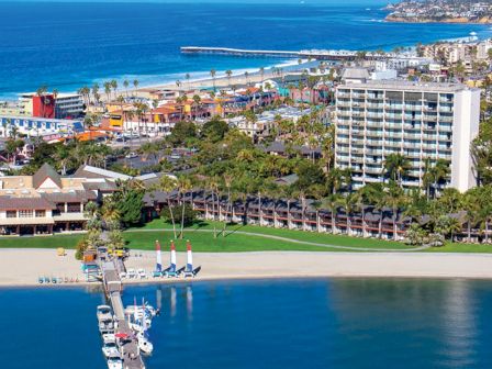 A sunny coastal resort with a marina, long pier, sandy beach, palm trees, and mid-rise hotels by turquoise water.