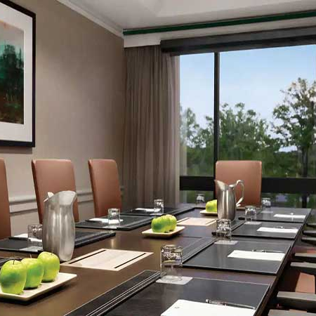 A modern conference room with a long table set for a meeting, pastel chairs, large window view, and polished place settings with green apples.