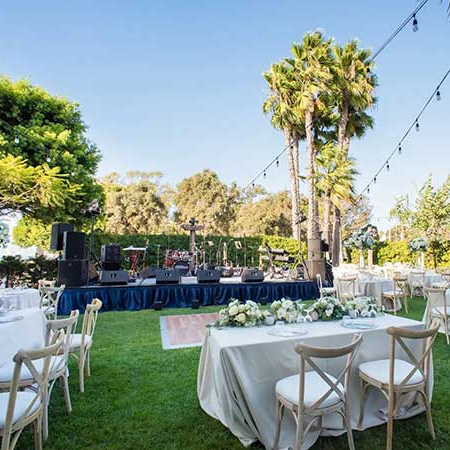 An outdoor wedding or event setup with round and rectangular tables, white linens, floral centerpieces, string lights, and a stage with sound equipment under a sunny, palm-tree filled garden.