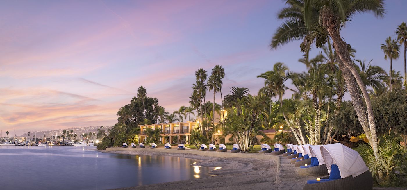 A tranquil beach at sunset with palm trees, calm waves, and row of umbrellas along the shore, a peaceful tropical getaway scene.