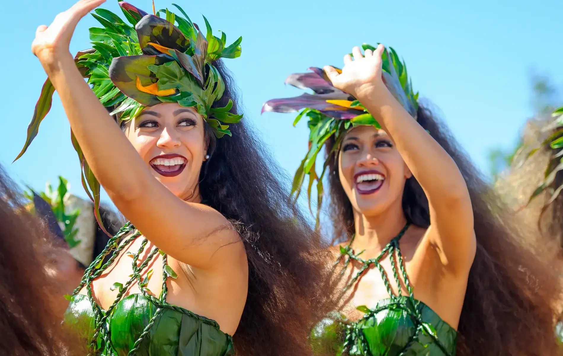 Two smiling women in green tropical outfits with leafy headdresses perform a dance, bright blue sky behind them.