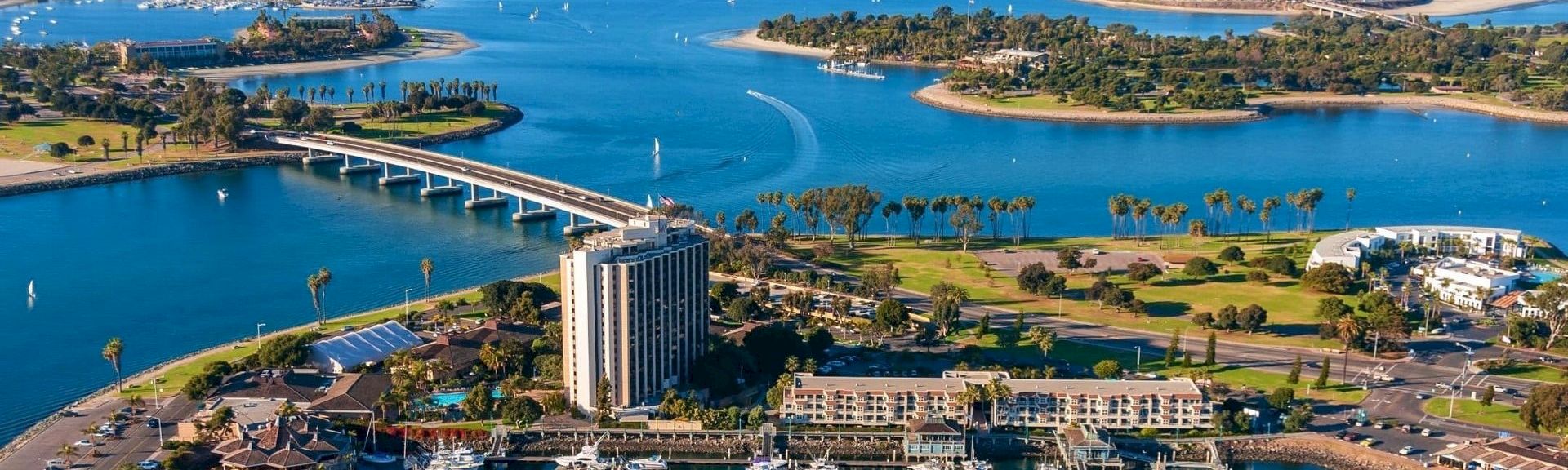 A sunny coastal city with a river inlet, palm trees, a tall hotel, a bridge, boats, and a marina along a scenic shoreline.