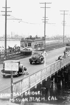 A black-and-white photo of Mission Bay Bridge in Mission Beach, CA, showing a busy road with cars, trucks, pedestrians, and a waterfront ledge; vintage signage and power poles line the span.