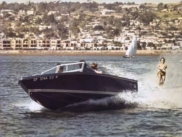 A person jetskiing behind a dark blue motorboat on calm water, with a coastal town and hills in the background.