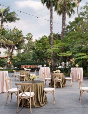 An outdoor event setup with round tables draped in blush and olive linens, string lights, palm trees, and elegant chairs.