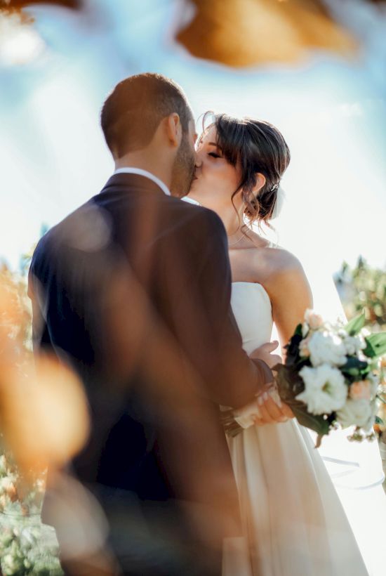 A bride and groom share a kiss at their wedding, framed by soft flowers and warm sunlight, creating a romantic, intimate moment.