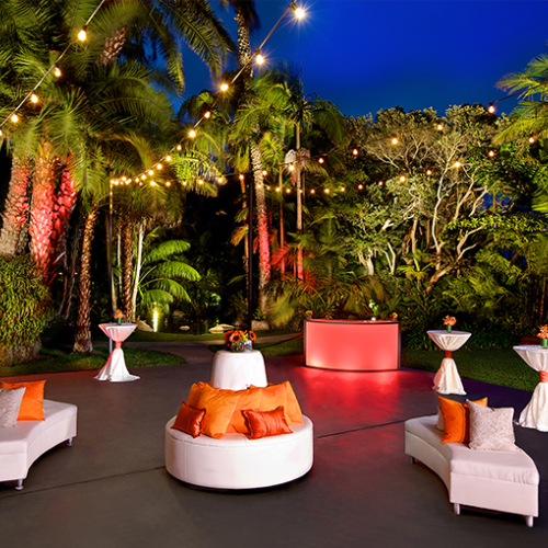 An outdoor lounge with white round seating, orange cushions, tropical plants, string lights, and a glowing red centerpiece under a blue evening sky.