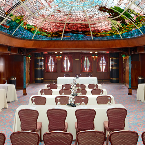 Elegant conference room set for a banquet with white-clothed tables, arranged chairs, and a colorful glass dome ceiling over spotless decor.