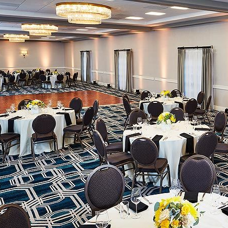 A banquet hall with round tables, yellow floral centerpieces, and chairs set for a formal event, warm lighting and a patterned carpet.