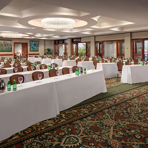 A conference room set for a banquet: long white-covered tables with green bottles, glassware, chairs, chandeliers, and artwork on the walls.