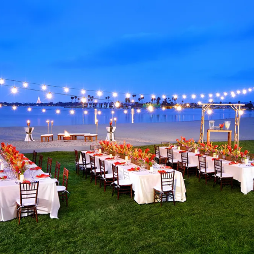 A lakeside evening wedding setup with long banquet tables, white linens, red floral centerpieces, string lights, and chairs on a grassy shore by the water.