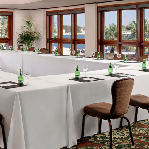 A conference-style dining room set up around a U-shaped table with white linens, green glass bottles, and brown chairs, by large windows overlooking water.