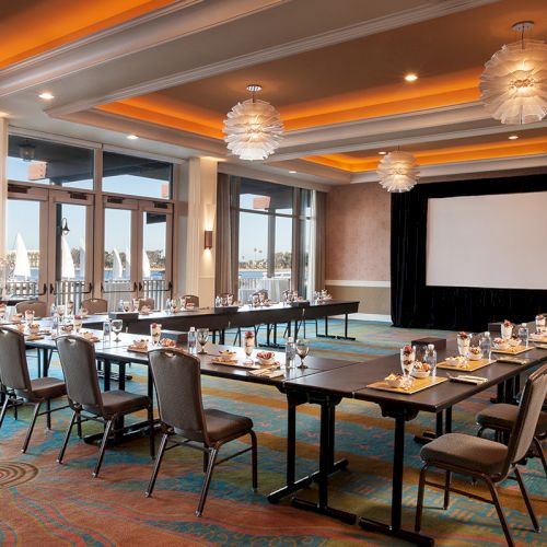 A stylish conference room setup with a U-shaped table, a large projector screen, floor-to-ceiling windows, and warm ambient lighting.