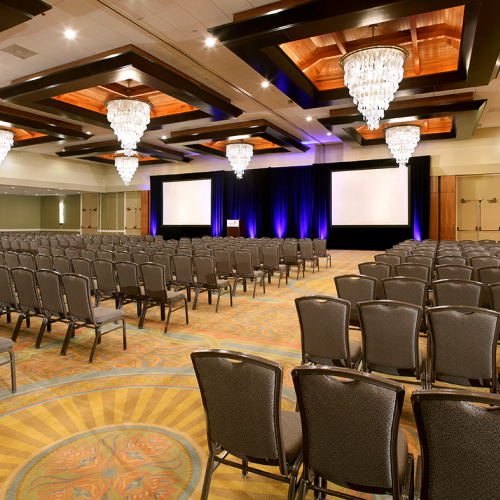 A large conference hall set for a presentation, with rows of chairs, a stage, and chandeliers, all under warm lighting.