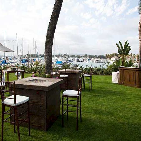 A seaside outdoor setup with wooden bar tables and stools on green grass, palm trees, and boats/harbor in the background.