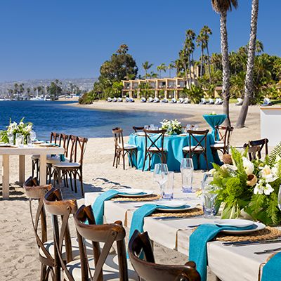 Outdoor beach wedding or banquet setup along the shore with tables, chairs, white and teal accents, floral centerpieces, and palm trees by the water.