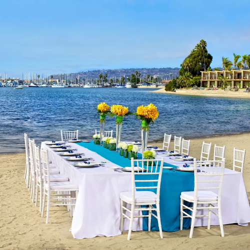 A beach wedding setup with a long white-draped table, turquoise runner, white chairs, yellow floral centerpieces, and calm harbor in the background.
