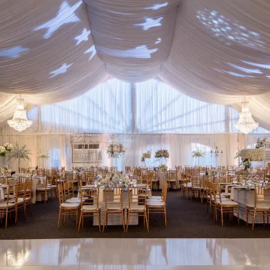 A lavish wedding reception setup under a draped tent with chandeliers, long banquet tables, gold chairs, and floral centerpieces.
