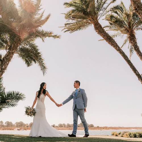 A bride and groom hold hands under tall palm trees by a sunny shoreline, wedding attire, serene outdoor ceremony vibe.