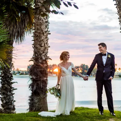 A bride and groom hold hands by a lakeside, surrounded by palm trees at sunset, smiling in wedding attire.
