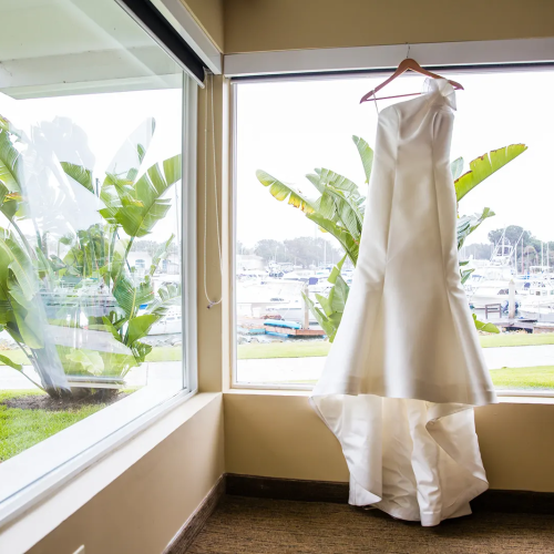 A white wedding dress hangs on a hanger by a window, with palm trees and a marina visible outside the glass walls, bright and serene.