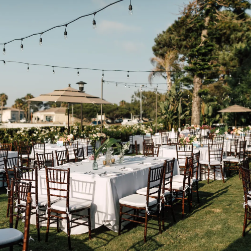 Outdoor wedding reception setup with long rows of round and rectangular tables, white linens, chiavari chairs, string lights, and green lawn scenery.