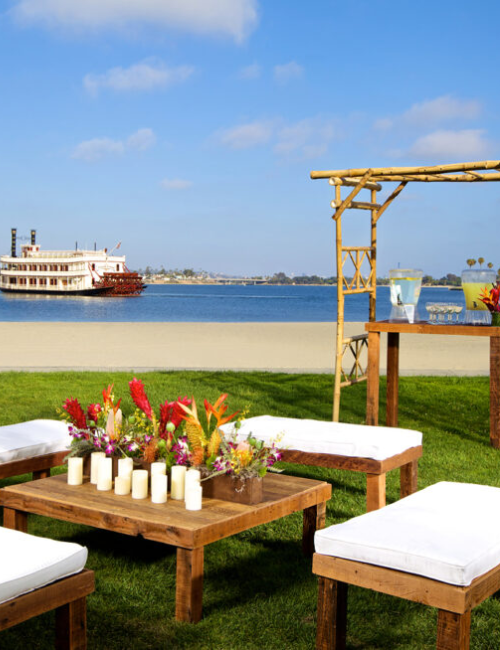 A lakeside outdoor setup with a wooden lounge area, four cushioned stools around a central coffee table, flowers, and a wooden arch by the water.