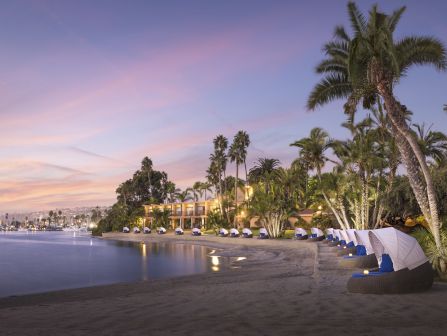 A tranquil beach at sunset with palm trees, calm water, lounge chairs, and thatched cabanas along the shore.
