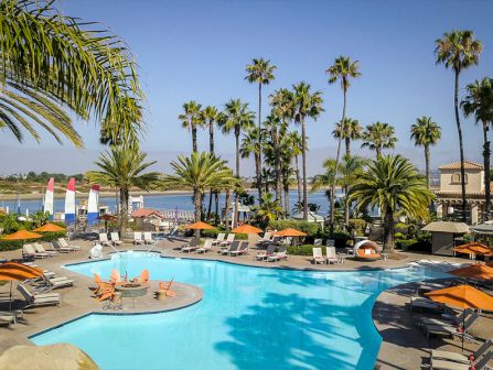 A sunny resort pool scene with palm trees, lounge chairs, and orange umbrellas by the water, oceanside.
