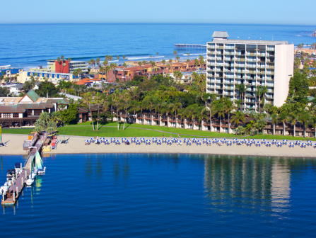 A resort beachfront with sun umbrellas, a pier, palm trees, a large hotel, and calm blue ocean water beyond the sandy shore.