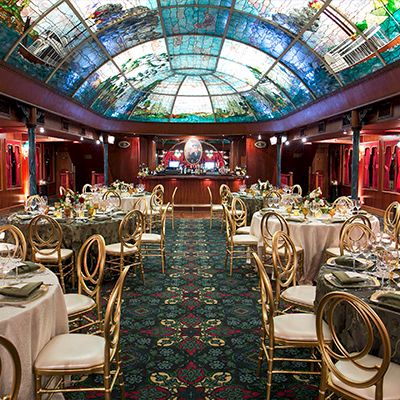 A luxurious banquet hall set with round tables, white chairs, fine china, floral centerpieces, and a stage at the back under a stained glass ceiling.