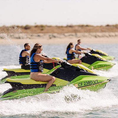 People riding green jet skis on water, wearing life vests, speeding past the shore. There&rsquo;s a sandy beach in the background.