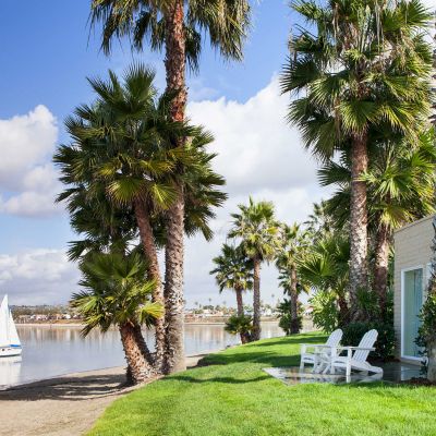A lakeside yard with palm trees, a green lawn, white chairs, a patio, and a small sailboat on calm water by a waterfront home.