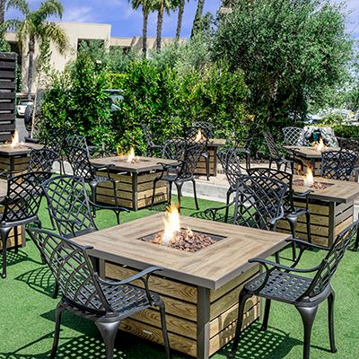An outdoor dining area with square tables, metal chairs, central fire pits, planters, and green lawn under sunny skies.