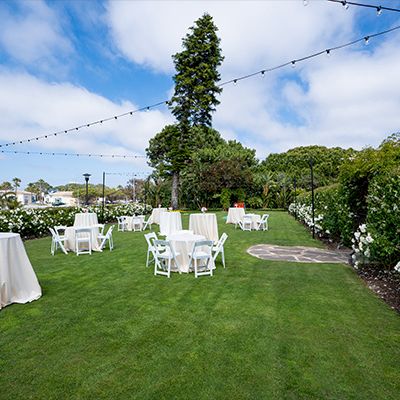 A well-kept outdoor garden setup with white round tables and chairs, string lights, and lush greenery under a blue sky, ready for a party.