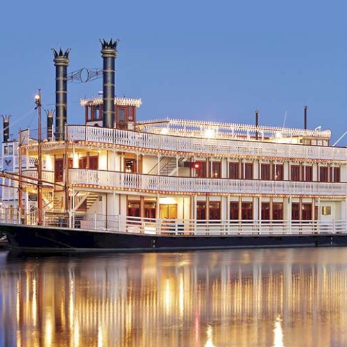 A grand, multi-deck river steamboat lighting up the water at dusk; tall smokestacks, paddlewheel, and warm reflections.