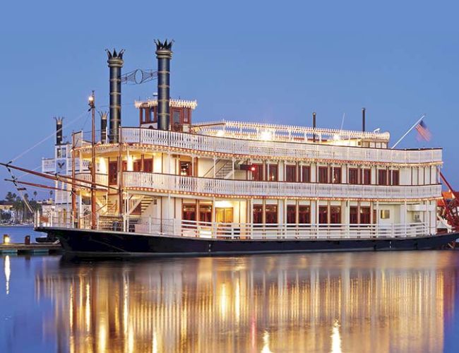 A grand, multi-deck river steamboat lighting up the water at dusk; tall smokestacks, paddlewheel, and warm reflections.