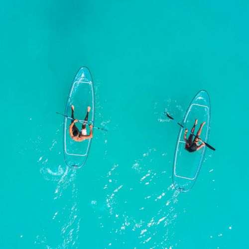 Two people stand on separate transparent surfboards riding turquoise water, paddling apart in calm, crystal-clear sea, enjoying sunny waves.