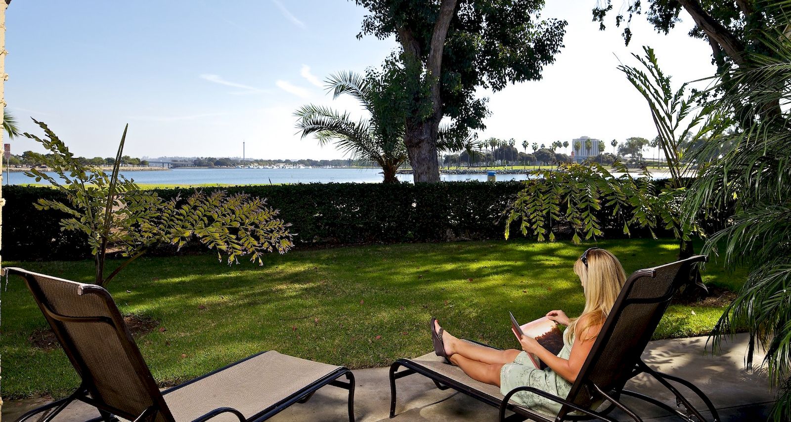 A woman lounging by a lawn, reading a book on a chair near a poolside with trees, water, and distant skyline in the background.
