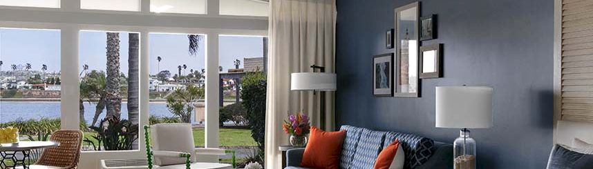 A bright hotel suite with a blue accent wall, large windows opening to a balcony, a bed, a blue sofa with orange pillows, and stylish lamps.