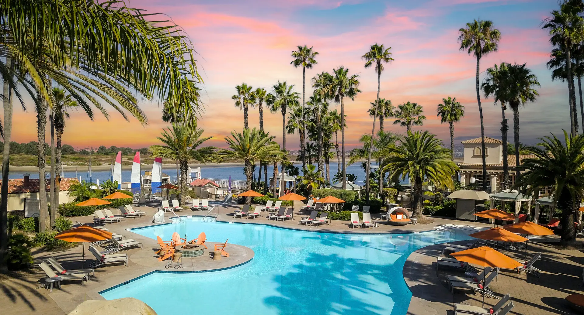 A tropical resort pool scene at sunset, with palm trees, lounge chairs, umbrellas, and a calm, blue pool reflecting the sky.
