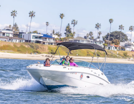 A white speedboat cruises on blue water near a sandy shore with palm trees and houses in the background, while three people enjoy the ride.